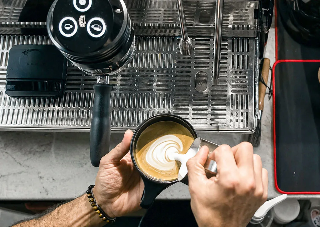 Overhead view of a Coffee Malem barista pouring latte art beside a professional espresso machine.